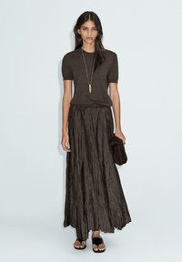 Haut en tricot marron à manches courtes associé à une longue jupe plissée marron. Le mannequin tient une pochette noire, portant des sandales noires.