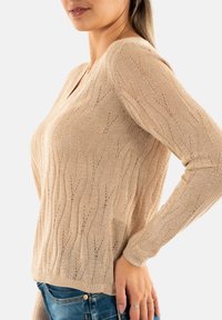 Maglione beige lavorato a maglia con scollatura a V, maniche lunghe e motivo ondulato. Texture morbida con dettagli a punto aperto e polsini e orlo a costine.