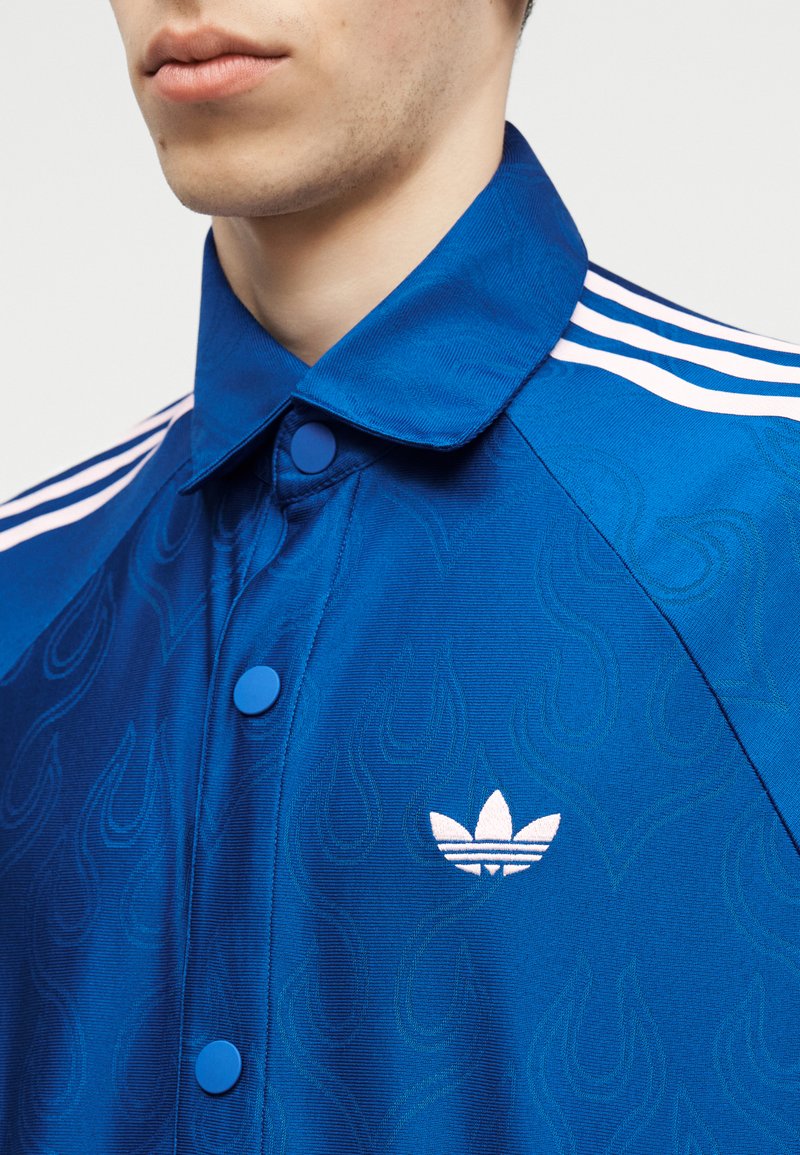 Primo piano di una persona che indossa una giacca Adidas blu con logo bianco, bottoni a pressione blu e strisce bianche sulla spalla.