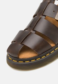 Ruskea nahkasandaali, jossa on kudottu yläosa, avoin varvas ja kontrastivärinen keltainen tikkaus mustassa kumipohjassa. Rakeinen pohja.