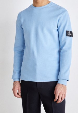 Lyseblå langærmet t-shirt lavet af tekstureret stof med rund halsudskæring og ribbede manchetter, der har et sort logo-patch på ærmet.