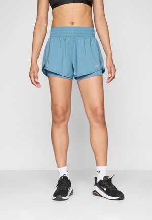 Kobieta ubrana w czarny sportowy biustonosz, jasnoniebieskie krótkie spodenki do biegania Nike, białe skarpetki Nike i czarne sneakersy Nike, stojąca na neutralnym tle.