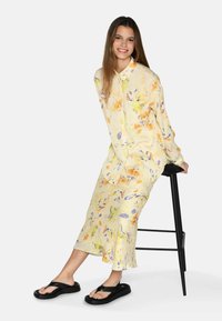 Abito floreale giallo con maniche lunghe, caratterizzato da una morbida trama e una silhouette fluida. Infradito neri completano l'outfit.