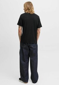 Schwarzes T-Shirt mit kurzen Ärmeln kombiniert mit dunkelblauen Weitbein-Jeans, die über Steppdetails und Gesäßtaschen verfügen. Sichtbare legere Schuhe.