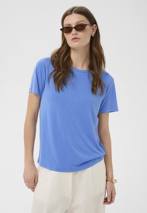 Femeie cu păr lung și castaniu, purtând ochelari de soare maro, cămașă albastră cu mâneci scurte, pantaloni albi, cercei aurii, brățară și inele pe un fundal alb.