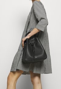 Svart läder bucket-väska med texturerad yta och snörstängning. Rundad botten, korta handtag och diskret branding på framsidan.