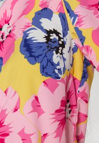 Blusa floral com flores grandes em rosa e azul sobre um fundo amarelo. O tecido parece suave, com cores vibrantes e padrões audaciosos.