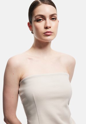 Młoda kobieta z zaczesanymi do tyłu włosami, nosząca srebrne kolczyki koła i beżowy top bez ramiączek na jednolitym białym tle.