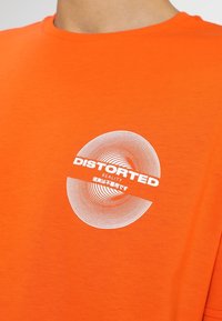 Oranžové bavlněné tričko s krátkým rukávem, které obsahuje kruhový grafický design s bílým textem "DISTORTED REALITY" a soustřednými liniemi.
