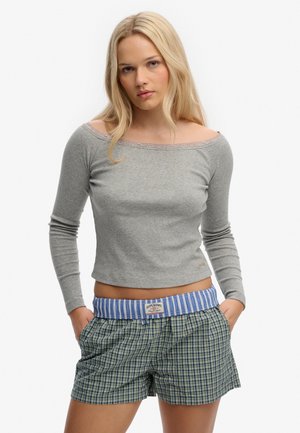 Blonde vrouw die een grijs off-shoulder top met lange mouwen en groene geruite korte broek met een blauwe gestreepte tailleband draagt, handen in de zakken.