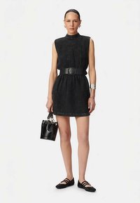 Mini robe noire sans manches avec un col montant, tissu texturé, taille cintrée avec une large ceinture, associée à un sac à main noir et des ballerines.