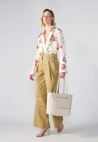 Blomsterbluse i off-white og pink, højtaljede beige bukser og en tekstureret creme farvet tote bag; model bærer nude hæle.