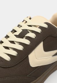 Braune Wildleder-Sneaker mit cremefarbenen Schnürsenkeln und cremefarbenem Logo-Akzent. Glatte Textur mit abgerundeter Zehenpartie und Gummisohlen-Detail.