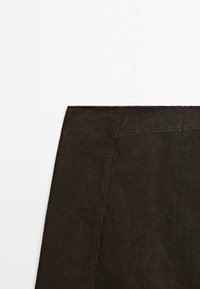 Tissu en velours côtelé marron foncé avec des côtes verticales montrant une partie de la section supérieure d'un vêtement et la couture de la ceinture sur fond blanc.