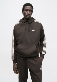 Sweatshirt castanha com bolso canguru, com três riscas brancas nas mangas e logótipo branco no peito; tecido macio e corte descontraído.