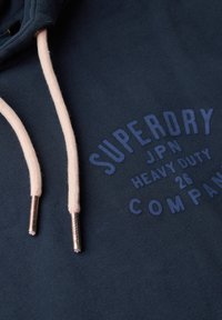 Sudadera marina hecha de una tela suave con un estampado de logo en azul oscuro. Los cordones son de color crema con puntas de metal.