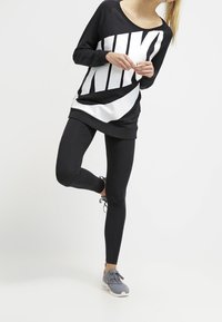 Svart sweatshirt med stor vit "NIKE"-logotyp, kombinerad med svarta leggings och grå sneakers. Avslappnad träningsklädsel med en luftig passform.