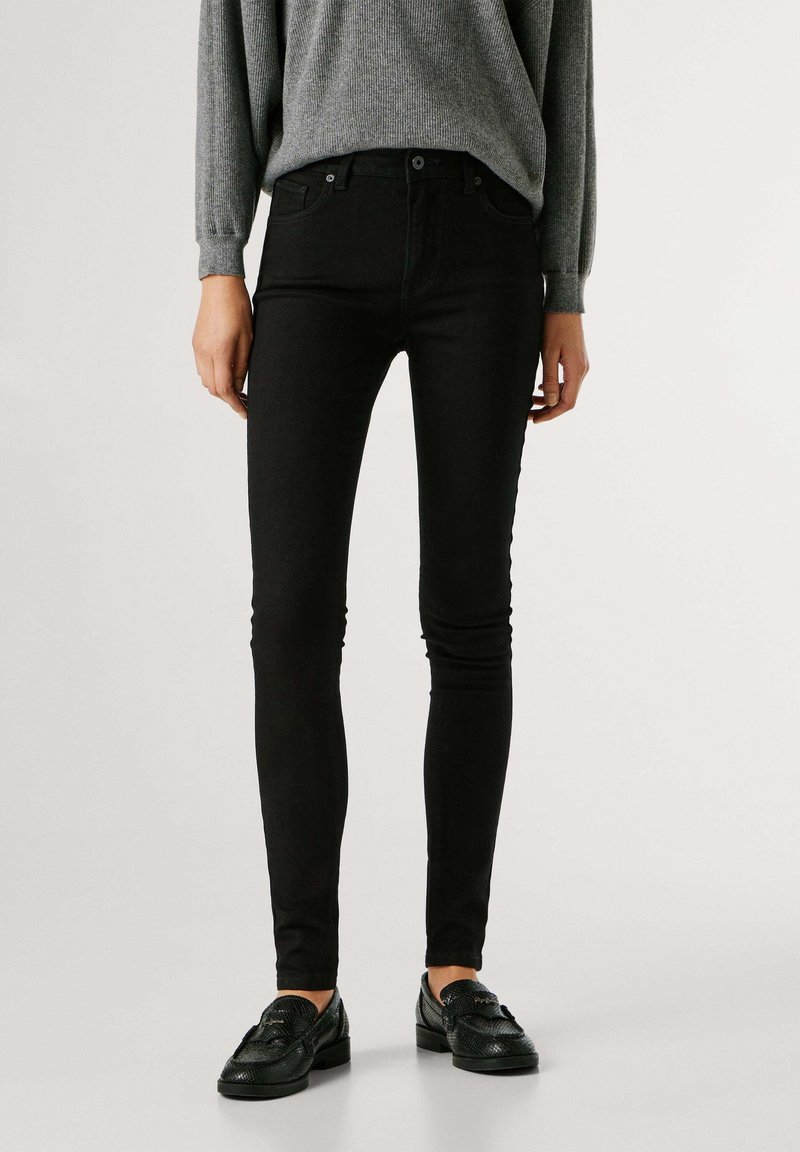 Juodi liekni džinsai iš medvilnės mišinio, su aukštu liemeniu, penkių kišenių dizainu ir metaline sagtele, derinami su pilku megztiniu ir juodais batais.