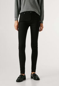 Jeans skinnies negros en mezcla de algodón, con una cintura alta, diseño de cinco bolsillos y cierre de botón metálico, combinados con un suéter gris y zapatos negros.