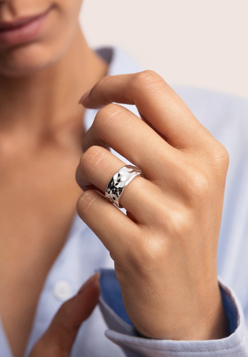 Anillo de plata esterlina con una superficie lisa y pulida, y acentos ondulados con textura, llevado en una mano ubicada cerca de una camisa azul claro.