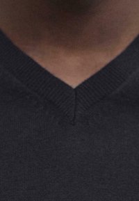 Pull en maille noire avec un col en V, présentant une surface texturée et un col côtelé. Le tissu semble doux et étroitement tissé.
