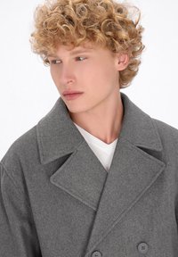 Cappotto grigio in misto lana con ampi risvolti, bottoni doppi e una texture morbida, indossato sopra una t-shirt bianca.