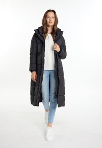 Cămașă puffer neagră cu un design lung, având cusături orizontale, închidere cu fermoar și glugă. Purtată peste un pulover alb și blugi deschiși.