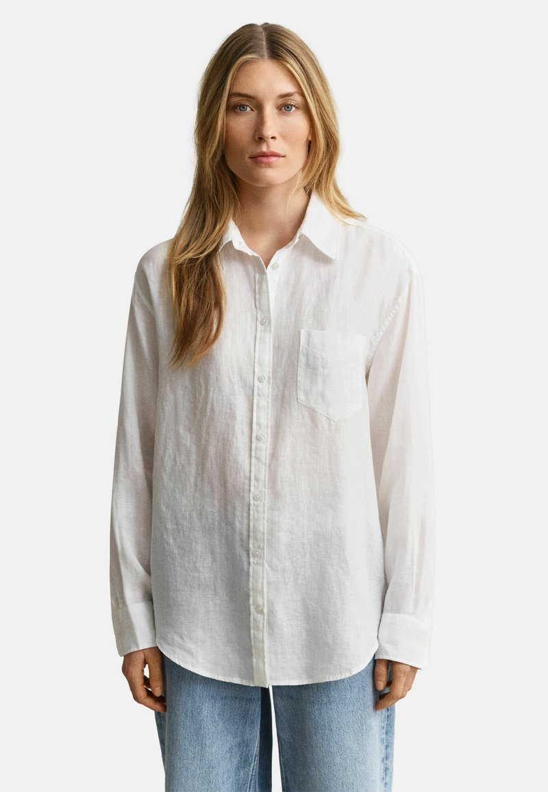 Mujer rubia con cabello largo, vestida con una camisa blanca abotonada con un bolsillo en el pecho y unos jeans azul claro, de pie contra un fondo blanco.