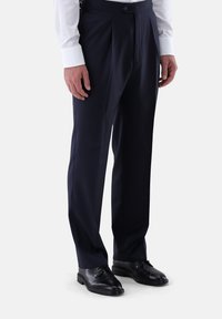 Mann, der formelle dunkelblaue Hosen, schwarze Lederschuhe und ein weißes Langarmhemd trägt, vor einem schlichten Hintergrund steht.