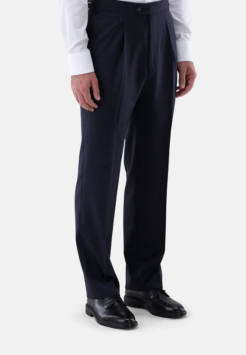 Mann, der formelle dunkelblaue Hosen, schwarze Lederschuhe und ein weißes Langarmhemd trägt, vor einem schlichten Hintergrund steht.