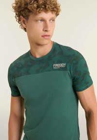 T-shirt vert à manches courtes avec un motif camouflage sur la partie supérieure, présentant une section lisse contrastante en dessous et une étiquette de marque.