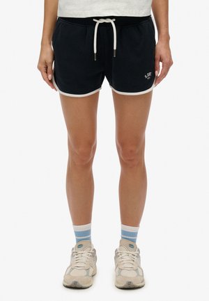 Zwarte katoenen korte broek met witte bies, elastische tailleband en trekkoord; logo gedrukt aan de linkerkant. Draagt lichtblauwe sportsokken en sneakers.