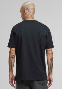 Juoda medvilninė marškinėliai su trumpomis rankovėmis, apvaliu kaklu ir laisvu siluetu. Nugarinėje pusėje yra sudėtingos tatuiruotės ant rankų ir kaklo.