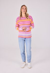 Gestreifter Pullover in mehreren Farben mit Farbtönen von Rosa, Orange und Lavendel; gestrickte Textur; kombiniert mit hellblauen Jeans und weißen Turnschuhen.