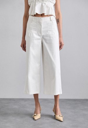 Witte verkorte wijde broek met geplooide voorkant, gemaakt van een gladde stof. Gecombineerd met beige hoge hakken.