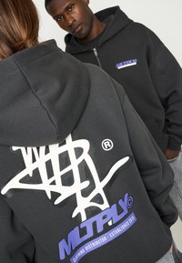 Zwarte hoodie met grote witte afbeelding op de achterkant, en een klein blauw logo op de linkervoorzijde. De stof lijkt zacht en heeft een relaxte pasvorm.
