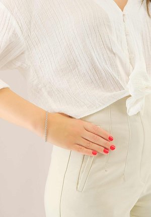 Blusa bianca con texture e dettaglio annodato, abbinata a pantaloni beige chiaro a vita alta. Un bracciale in argento adorna una mano con unghie rosse.