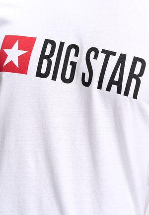 Balts kokvilnas T-krekls ar lielu sarkanu kvadrātu, kurā ir balta zvaigzne un trekns melns teksts "LIELĀ ZVAIGZNE" uz priekšpuses.