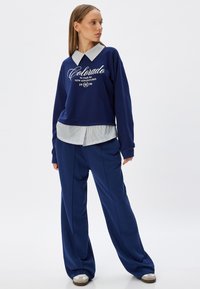 Sudadera navy con texto bordado en blanco que dice "Colorado" y cuello a rayas, combinada con pantalones navy holgados y zapatillas grises. Diseño sencillo.