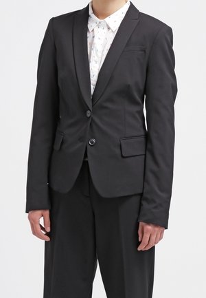 Černý přizpůsobený blazer se dvěma knoflíky, klopovými límci a klopovými kapsami. Nositý přes košili s límečkem a jemným vzorem. Hladká textura látky.