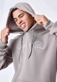 Sudadera gris claro hecha de un tejido suave, con un bolsillo frontal y texto bordado en blanco "Project + Paris" en el pecho.