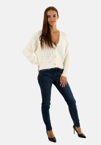 Cardigan bianco e soffice con scollo a V profondo e tre grandi bottoni, abbinato a jeans skinny blu scuro e scarpe nere con tacco alto.