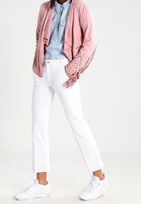 Lichtroze franje cardigan met studaccenten, gelaagd over een lichtblauw denim shirt. Gecombineerd met witte rechte broek en witte sneakers.