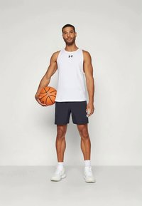 Atleet seisab, hoides paremas käes korvpalli, seljas valge ilma varrukateta särk, mustad lühikesed püksid, valged sokid ja valged tossud.