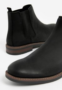 Sorte Chelsea-støvler i læder med elastiske sidepaneler, runde tæer og slidstærke gummisåler, med brun syet kant og afdæmpet tekstur.