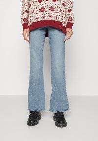 Jeans de ganga azul claro, com corte flare e um ligeiro desbotado. Combinados com sapatos pretos com detalhe de franjas e um pulôver de malha padrão em bege e vermelho.