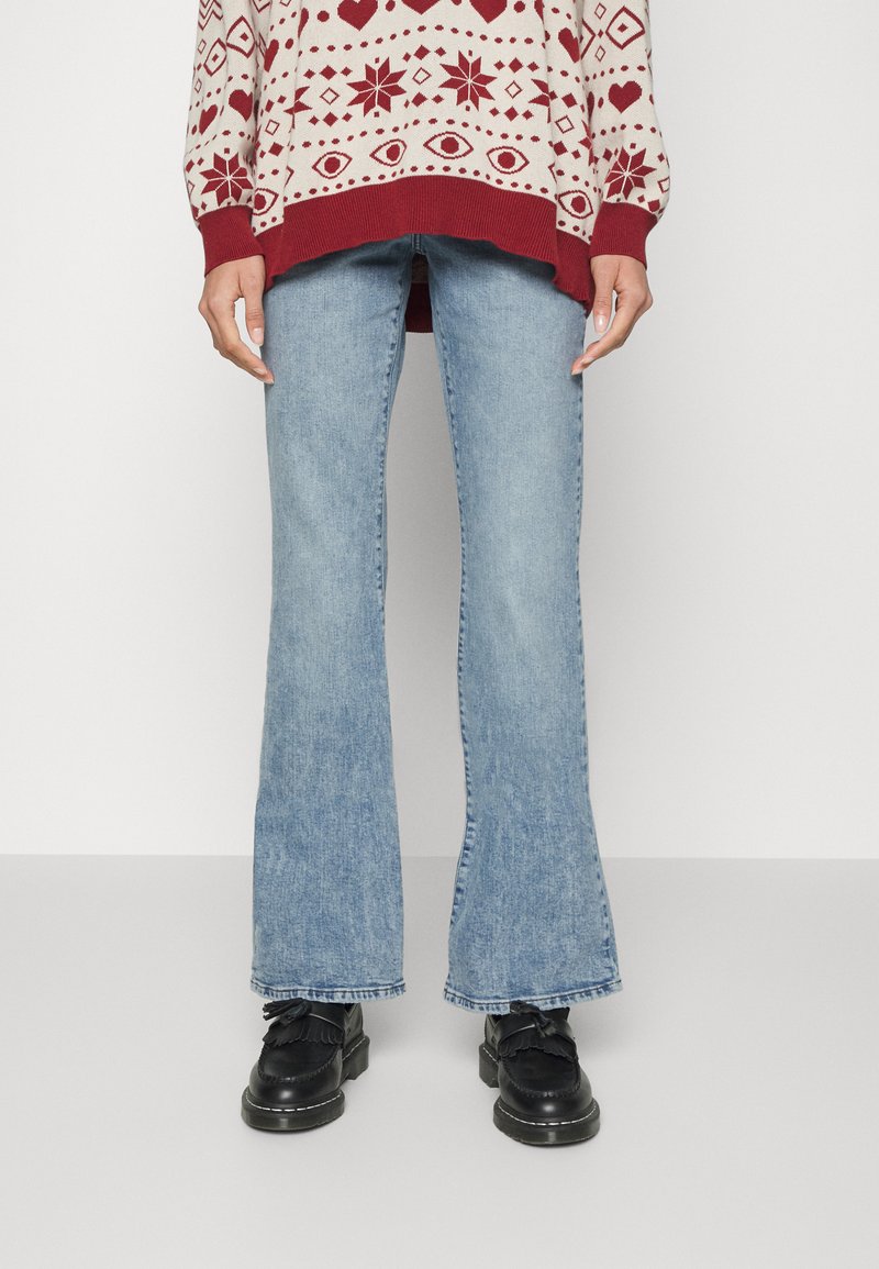 Jeans de ganga azul claro, com corte flare e um ligeiro desbotado. Combinados com sapatos pretos com detalhe de franjas e um pulôver de malha padrão em bege e vermelho.