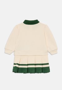 Robe longue à manches longues crème avec col vert foncé et jupe plissée, présentant deux rayures crème et une rayure verte horizontales près de l'ourlet.