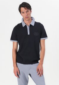 Camisa polo preta com um padrão texturizado, colarinho e punhos cinzentos, apresentando um patch de logotipo no peito. Tecido suave com mangas curtas.