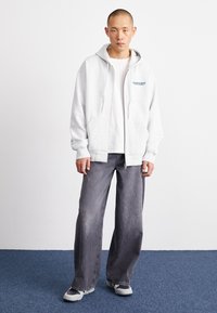 Grey zip-up hoodie with a front pocket and blue logo, worn over a white t-shirt. Paired with grey wide-leg jeans and grey sneakers.